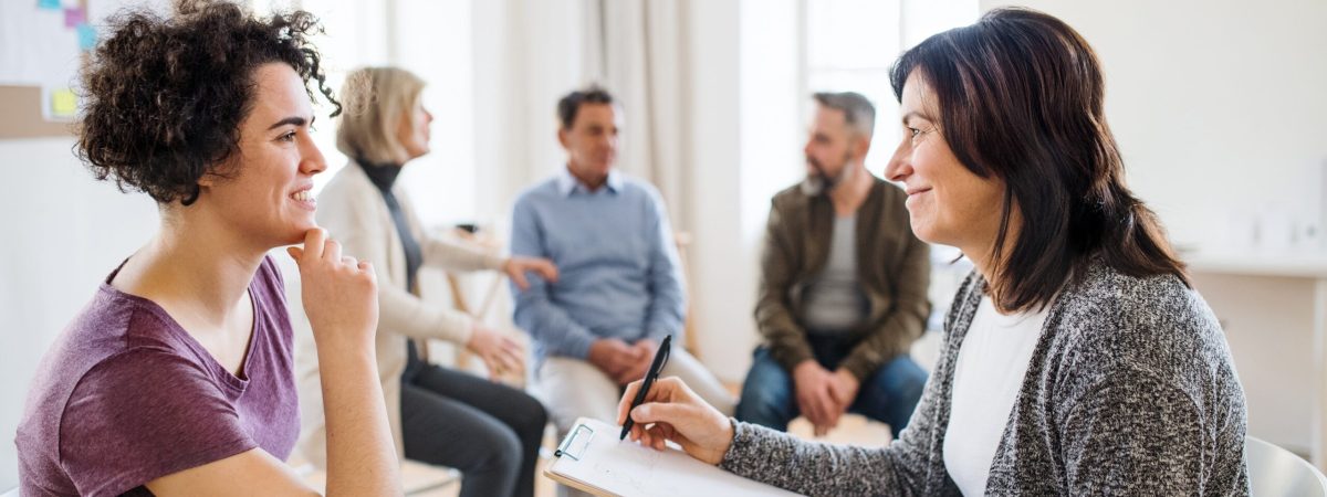 A senior counsellor with clipboard talking to a young woman during group therapy.